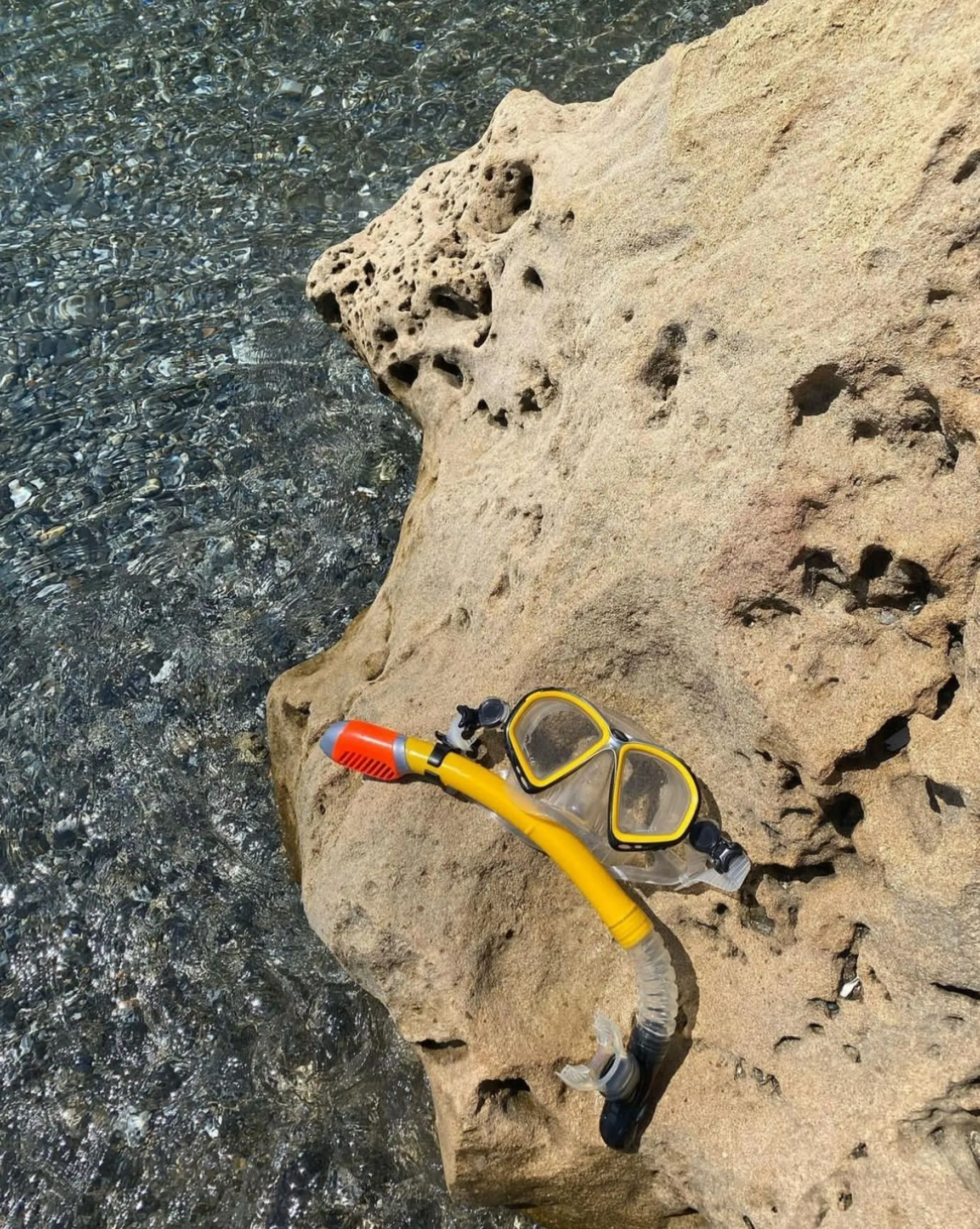 Eine gelbe Schnorchelmaske mit Schnorchel liegt auf einem Felsen neben klarem Wasser.